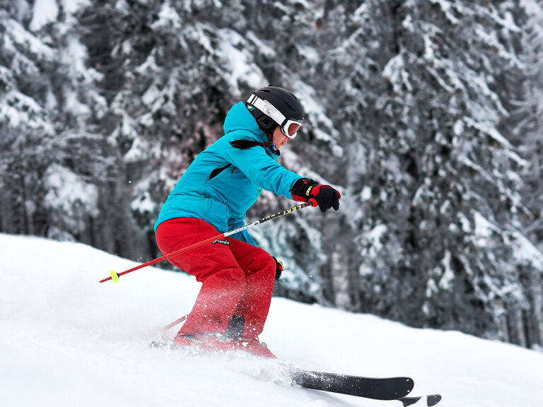 Eine Skifahrerin fährt durch frischen Pulverschnee vor verschneitem Wald.